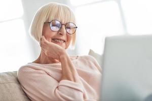 Focusing on something. Beautiful senior woman using laptop and smiling while relaxing on the couch at home photo