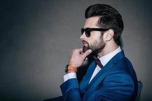 Gorgeous and handsome. Close-up side view of young handsome man in suit and bow tie holding hand on chin and looking away while sitting against grey background photo