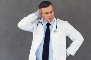 Hard to diagnose. Mature male doctor scratching his head and looking away while standing against grey background photo