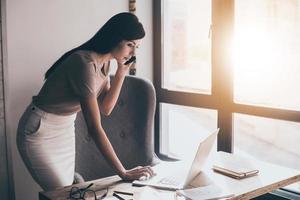 Let me check my timetable Side view of young beautiful woman talking on mobile phone and using her laptop while standing at her working place photo