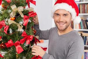 Decorating a Christmas tree. Cheerful young man decorating a Christmas tree and smiling at camera photo