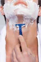 Close up of shaving. Close up of young man shaving his face while standing against grey background photo