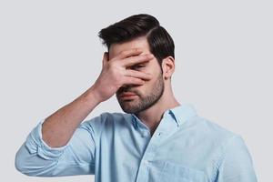 What is there Handsome young man covering eyes with hand and peeking while standing grey background photo