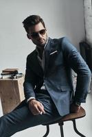 Looking just perfect. Charming young man in full suit looking away and holding a cigarette while sitting on the stool photo