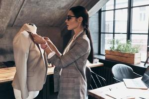 Everything must be perfect. Serious young woman in eyewear adjusting a collar of the jacket on mannequin while standing in her workshop photo
