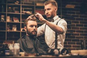 Making haircut look perfect. Young bearded man getting haircut by hairdresser while sitting in chair at barbershop photo
