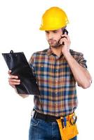 Concentrated on work. Confident young male carpenter in hardhat talking on mobile phone and looking at his clipboard while standing against white background photo