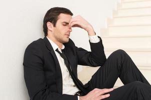 Tired and depressed. Side view of depressed young man in formalwear touching his nose and keeping eyes closed while sitting on staircase photo