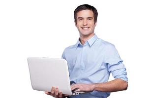 chico guapo de la computadora. un joven apuesto con camisa azul sosteniendo una laptop y sonriendo mientras se encuentra aislado en blanco foto