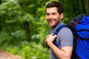 guapo turista. un joven apuesto que lleva una mochila y mira por encima del hombro con una sonrisa mientras está de pie en la naturaleza foto