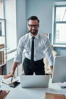 Handsome young man in formalwear looking at camera and smiling while working in the office photo