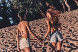 mejores amigos. dos mujeres jóvenes atractivas en pantalones cortos y trajes de baño tomados de la mano mientras corren en la playa foto