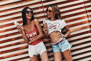 chicas con estilo. dos atractivas mujeres jóvenes sonriendo y sosteniendo helado mientras están de pie contra la pared de madera al aire libre foto