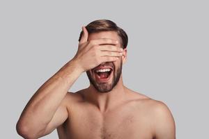 Can I look now Handsome young man covering eyes with hand and keeping mouth open while standing against grey background photo