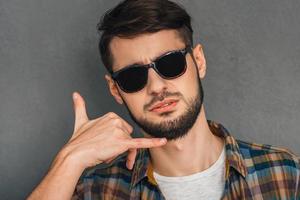 Call me Portrait of confused young man gesturing telephone with his hand and looking at camera while standing against grey background photo