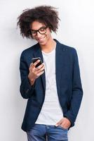 Typing message to friend. Handsome young African man holding mobile phone and smiling while standing against grey background photo