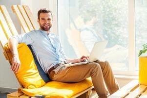 Ready to do business. Happy young man working on laptop and looking at camera while sitting inthe rest area of the office photo