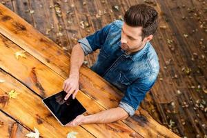 Enjoying his work outdoors. Top view of confident young man working on digital tablet while sitting at the wooden table outdoors photo