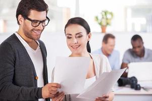 Working moments. Two cheerful business people holding documents and smiling while two people working on background photo