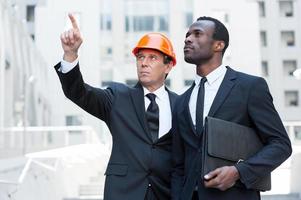 Discussing building project. Confident contractor in hardhat pointing away while standing together with African businessman photo