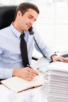 Busy working. Confident young man in formalwear making notes in his note pad and talking on the telephone while sitting at his working place photo