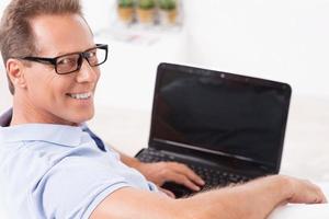 I am always online. Top view of cheerful mature man working on laptop and looking over shoulder while sitting on the couch at home photo