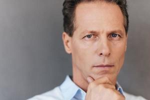 Portrait of confidence. Portrait of confident mature man holding hand on chin and looking at camera while standing against grey background photo