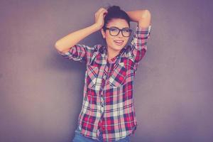 disfrutando de su estilo casual. hermosa joven mujer funky ajustando su cabello y sonriendo mientras está de pie contra el fondo gris foto