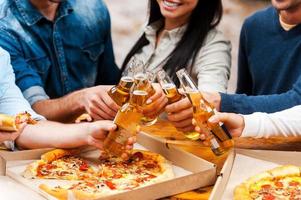 celebrando su amistad. primer plano de un grupo de jóvenes comiendo pizza y tintineando vasos con cerveza mientras están de pie al aire libre foto