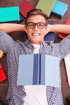 Happy bookworm. Top view of happy young man holding hands behind head and smiling while lying on the hardwood floor with colorful books laying all around him photo