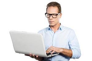 experto en ti en el trabajo. hombre maduro feliz en camisa sosteniendo una computadora portátil y ajustando sus anteojos mientras está de pie contra el fondo blanco foto