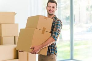 Moving to a new apartment. Cheerful young man holding a cardboard boxes and smiling at camera while other carton boxes laying on background photo