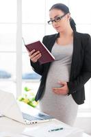 Pregnant woman reading book. Beautiful pregnant businesswoman reading a book while standing near her working place in office photo