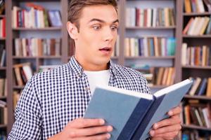 leyendo un libro emocionante. sorprendido joven leyendo un libro y manteniendo la boca abierta mientras está de pie en la biblioteca foto