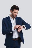 What time is it Handsome young man in full suit checking the time while standing against grey background photo