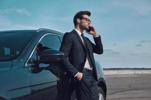 siempre disponible. apuesto joven hombre de negocios hablando por teléfono mientras está de pie cerca de su coche al aire libre foto