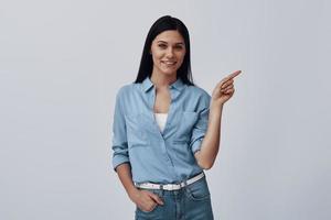 Attractive young woman looking at camera and pointing copy space while standing against grey background photo