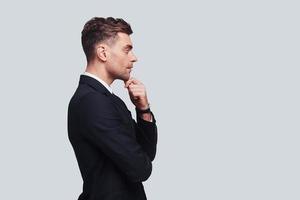 Taking time to think. Serious young man in full suit keeping hand on chin and looking away while standing against grey background photo