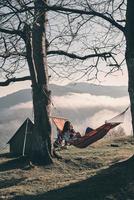 Starting a day with a blank page. Attractive young woman lying in hammock and holding a mug while camping in mountains photo