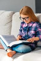 capítulo interesante. hermosa niñita con gafas leyendo un libro mientras se sienta en el sofá en posición de loto en casa foto