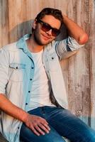 Carefree handsome. Handsome young man in sunglasses holding hand in hair and smiling while sitting against the wooden wall photo