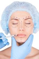 Botox injection in lips. Beautiful young woman in medical headwear and sketches on face keeping eyes closed while doctors hand making an injection in lips photo