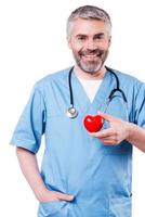 Cardiology surgeon. Confident mature cardiology surgeon holding heart shape toy and smiling while standing isolated on white photo