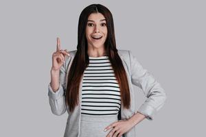 Great idea Gorgeous young woman in smart casual wear keeping finger raised and looking at camera with smile while standing against grey background photo