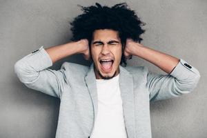 Stop this sound Frustratedyoung African man covering ears with his hands and keeping mouth open while standing against grey background photo