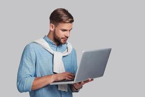 Concentrated at work.  Good looking young man working on his laptop while standing against grey background photo