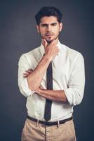 I will think about it. Confident young handsome man keeping hand on chin and looking at camera while standing against grey background photo