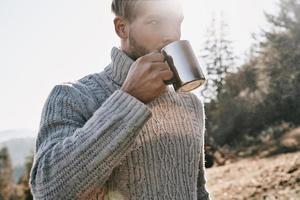 Fresh coffee tastes delicious. Handsome young man in warm sweater having morning coffee while camping in mountains photo
