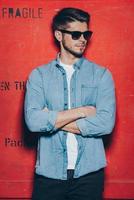 Stylish as always.Handsome young man in sunglasses keeping arms crossed and looking away while standing against red background photo