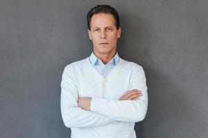 Confident mature man. Confident mature man keeping arms crossed and looking at camera while standing against grey background photo